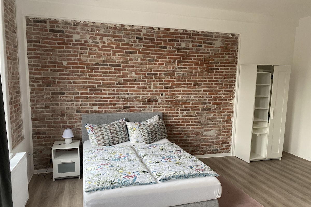 Quarto com parede de tijolos, cama com roupa de cama florida, mesa de cabeceira com abajur e guarda-roupa.