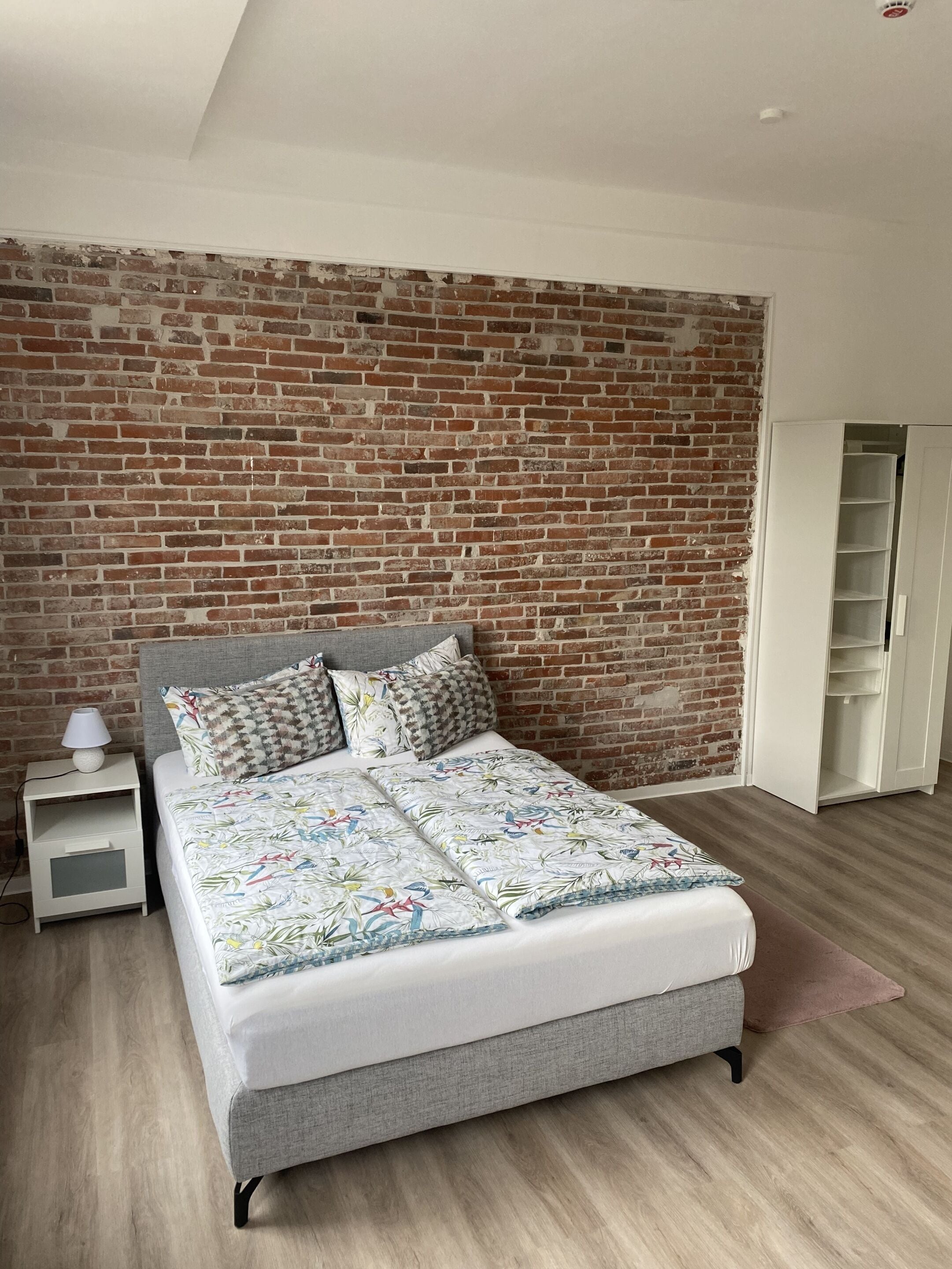Una camera da letto con parete in mattoni, un letto con copriletto decorato, un comodino con lampada e un armadio.