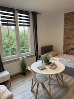 A modern bedroom with a large window, a bed with floral bedding, a round dining table with chairs, and a radiator.