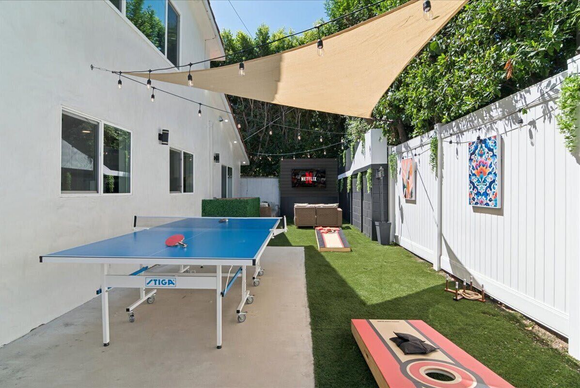 Backyard during the day with outdoor aluminum Stiga ping-pong table, corn hole, and large TV with sofa area for hanging out