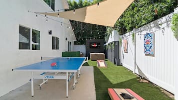 Backyard during the day with outdoor aluminum Stiga ping-pong table, corn hole, and large TV with sofa area for hanging out