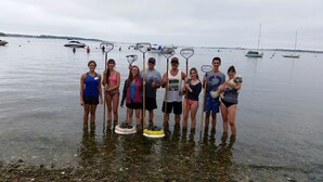 Eine Gruppe von Menschen steht im flachen Wasser mit Stand-Up-Paddles, ein ruhiger See und Boote im Hintergrund.