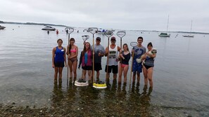 Eine Gruppe von Menschen steht im flachen Wasser mit Stand-Up-Paddles, ein ruhiger See und Boote im Hintergrund.