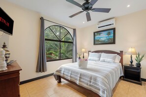 Habitación de hotel con cama, ventilador de techo, ventana con cortinas, un cuadro, mesita de noche con lámpara y televisor en la pared.