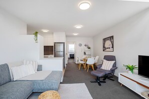 Una sala moderna con sofá, sillón y mueble para televisor. Cuenta con un comedor con mesa y sillas, además de una cocina con refrigerador.