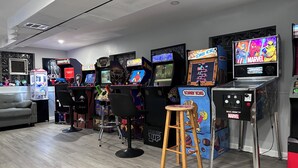 Una fila di cabinati arcade, tra cui giochi come 'Donkey Kong' e 'Marvel', in una stanza con un divano grigio e uno sgabello di legno.