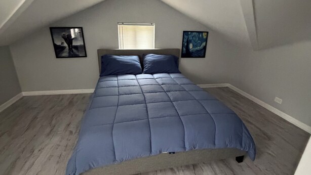 Una camera da letto con soffitto spiovente, un letto con copripiumino blu, due quadri appesi alle pareti e una finestra con persiane.
