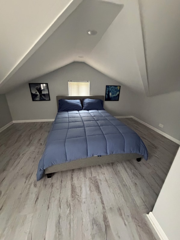 A bedroom with a sloped ceiling, a bed with a blue bedspread, two wall-mounted picture frames, and a window with blinds.