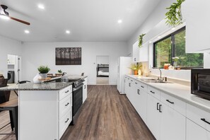 A modern kitchen with white cabinetry, a granite countertop, and a view of a bedroom through an open door. - Hot Tub Retreat on 190-Acre Farm · Sleeps 6 (Lakewood)