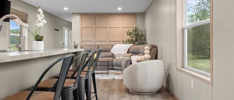 A modern kitchen with a dining area, built-in wooden cabinets, and a large window offering a view of the outdoors.
