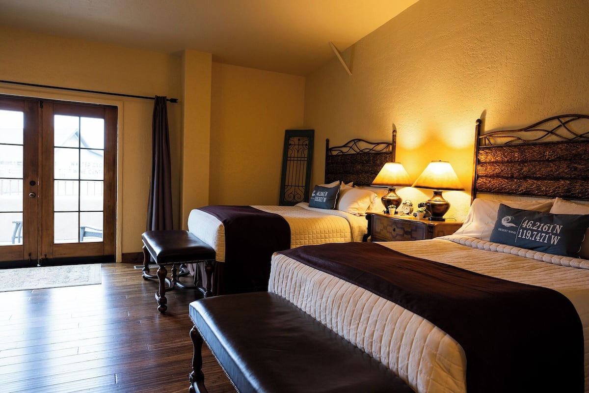 Habitación de hotel con dos camas, un banco, una mesita de noche con lámparas y vistas a través de un ventanal.