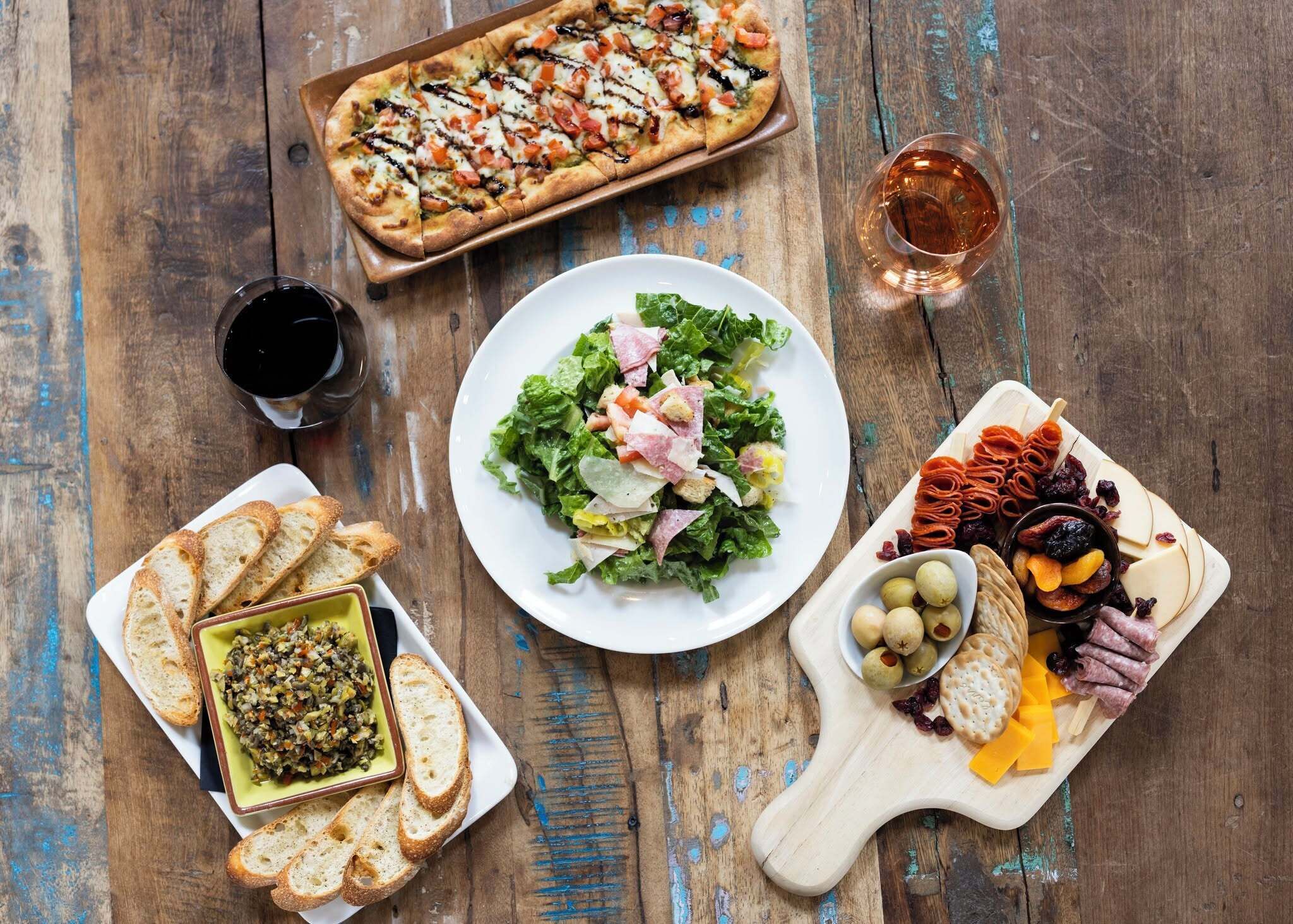 A wooden table with a variety of food items including a pizza, salad, bread, and a cheese board with meats and olives.