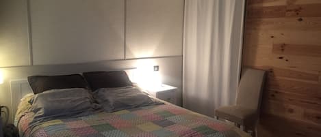 A bedroom with a bed, a bedside lamp, an air conditioner, a wooden wall panel, and a curtain.