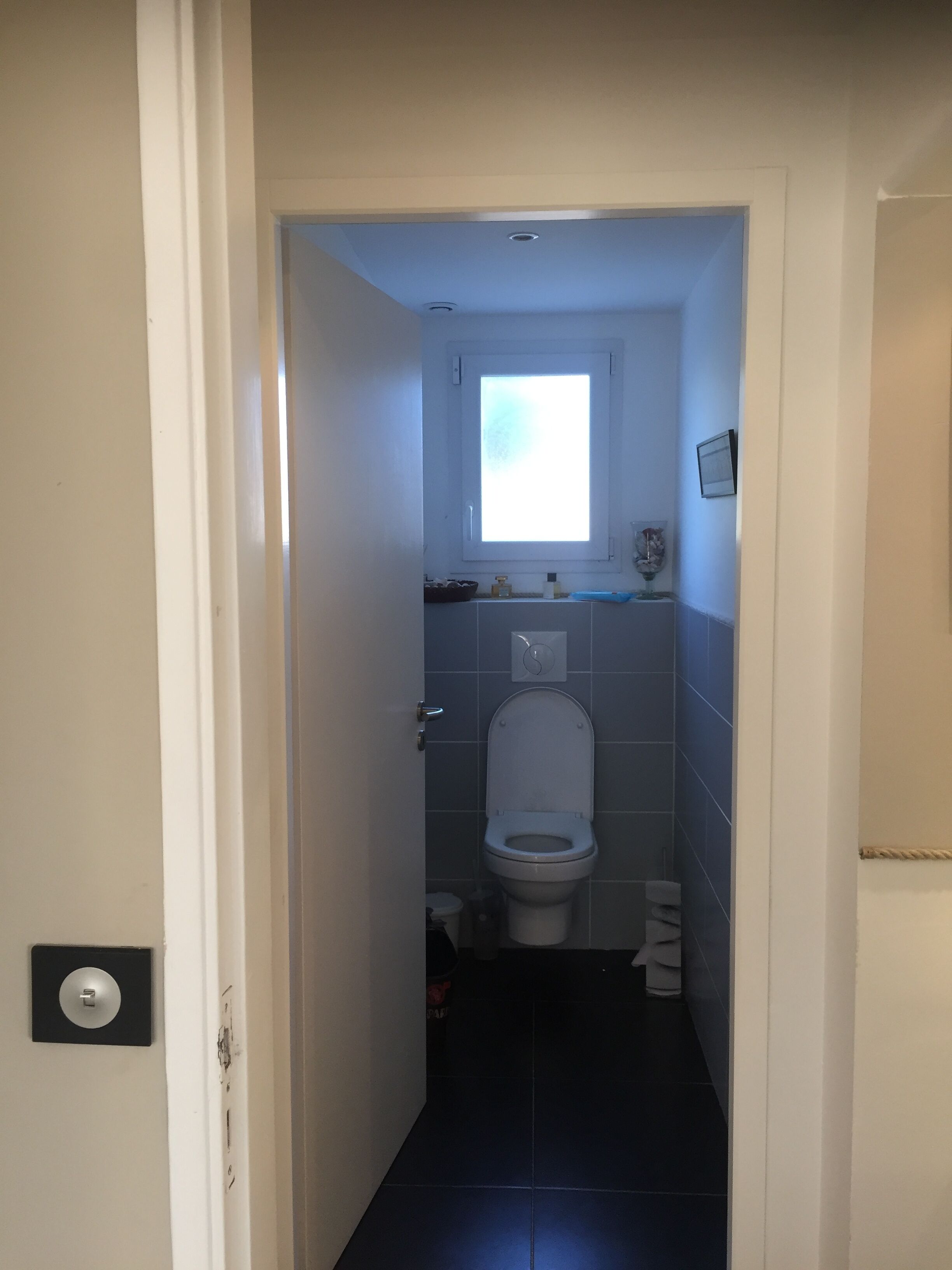 Ein schmaler Flur führt zu einem Badezimmer mit Toilette, einem kleinen Fenster und einer Wandleuchte.