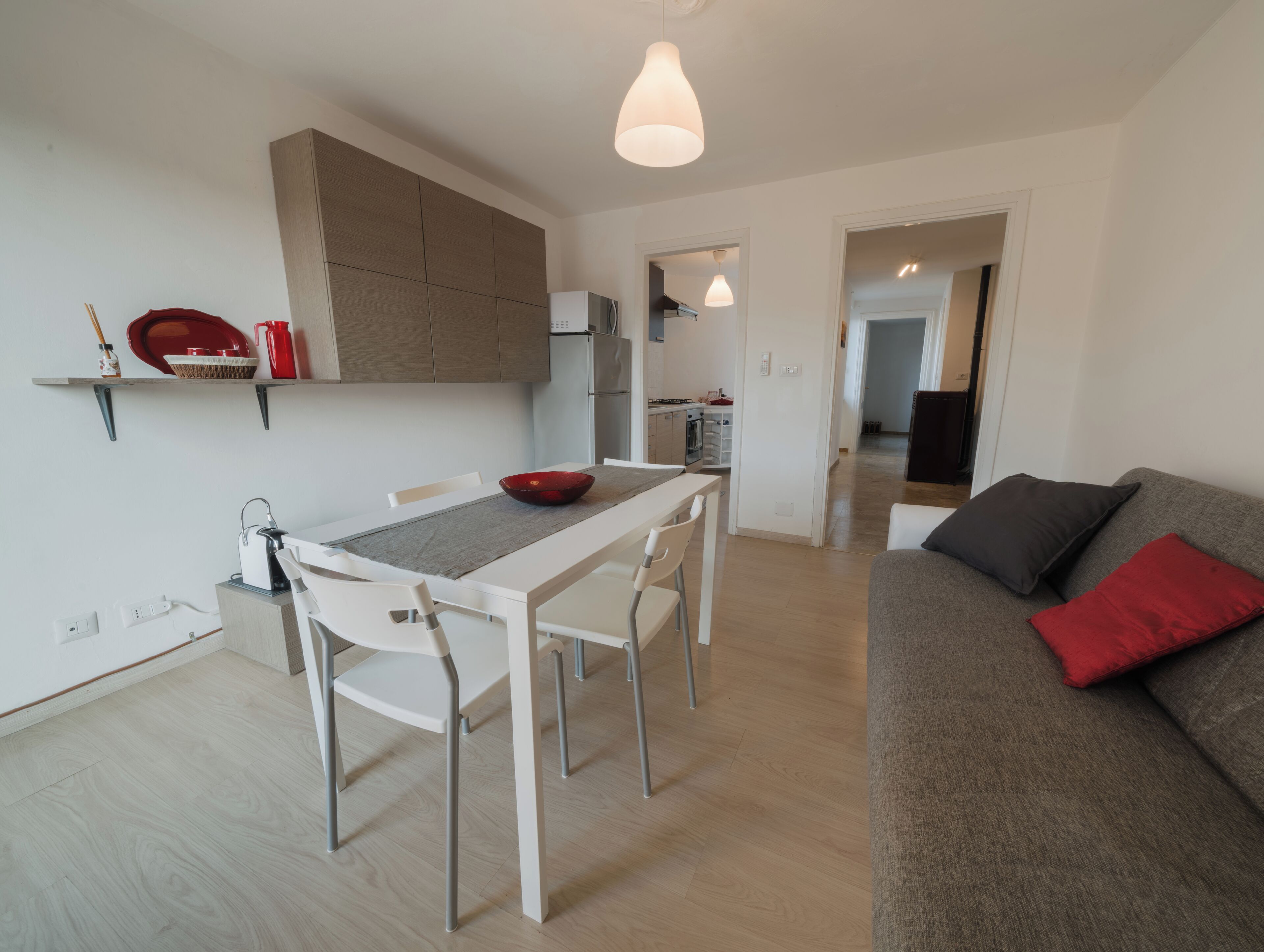 A modern kitchen with white cabinetry, a dining table with chairs, and a grey sofa with red cushions.