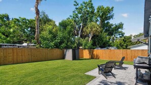 A backyard with a wooden fence, a grassy lawn, and a barbecue area with chairs. - Modern Hacienda | 3BR, Fenced Yard, Beach Gear, Dog-Friendly, Near Downtown (Sarasota)
