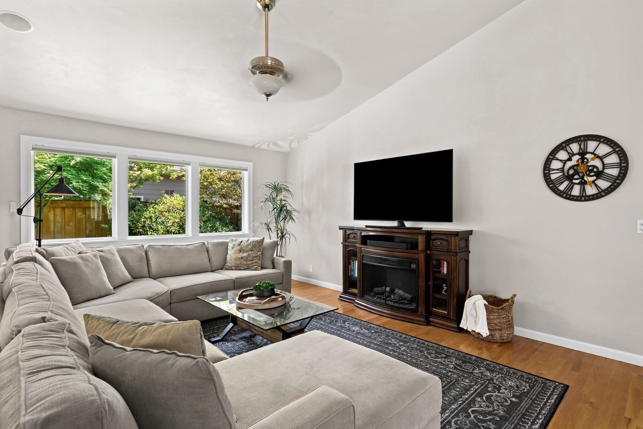 A living room with a sectional sofa, a flat-screen TV mounted on the wall, a fireplace, and a large window with a view of greenery.