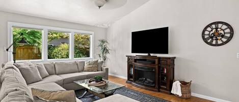A living room with a sectional sofa, a flat-screen TV mounted on the wall, a fireplace, and a large window with a view of greenery.