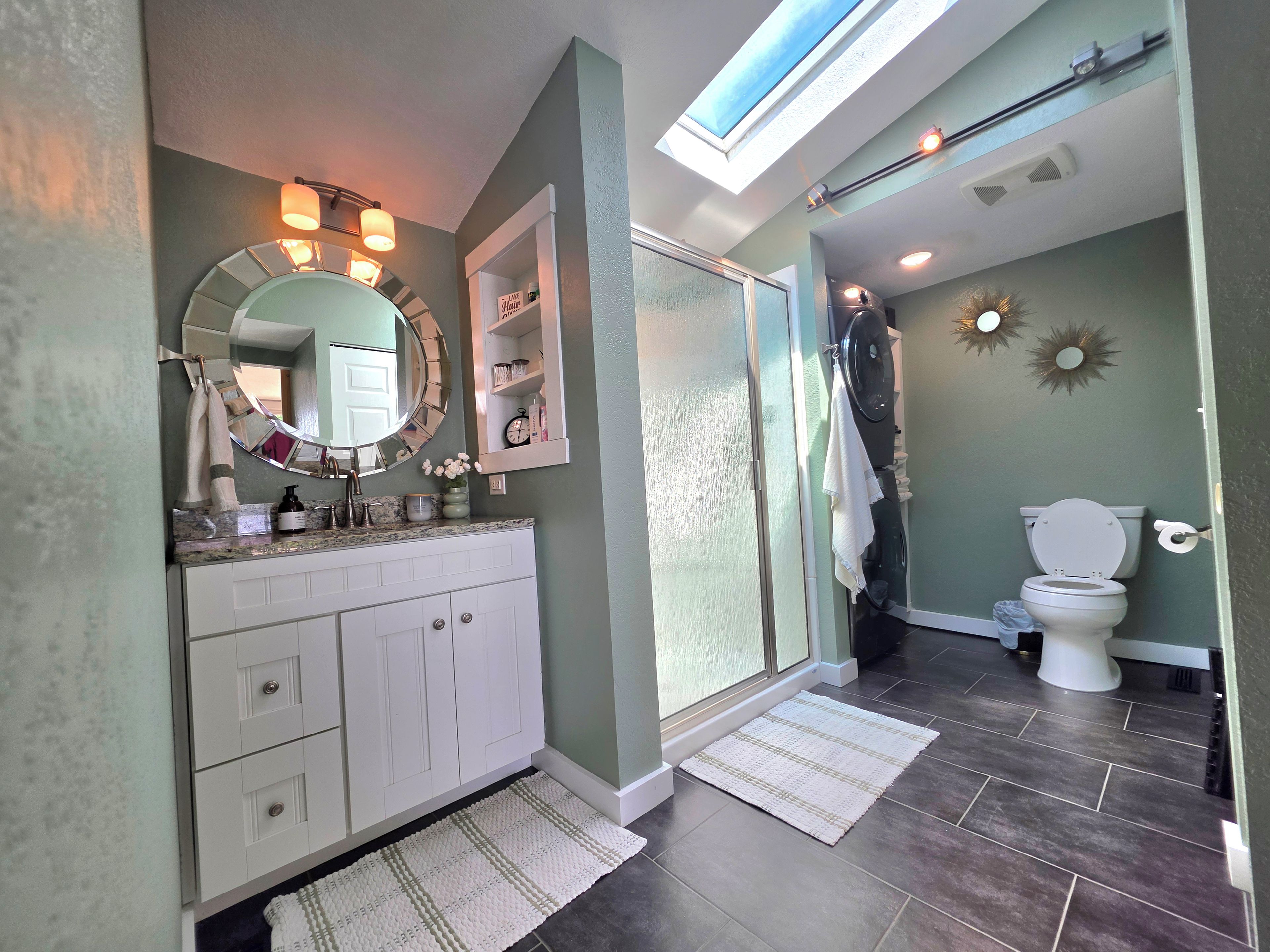 A bathroom with a vanity, mirror, towel rack, shower, toilet, and a skylight.