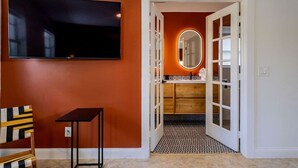 A room with a television mounted on the wall, a chair, a small table, and a doorway leading to a bathroom with a mirror and a sink. - 2-Acre Estate: Private Padel Court + Par-3 Golf (Davie)