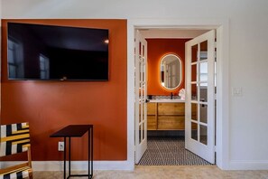 Ein Zimmer mit an der Wand befestigtem Fernseher, einem Sessel, einem kleinen Tisch und einer Tür zu einem Badezimmer mit Spiegel und Waschbecken.