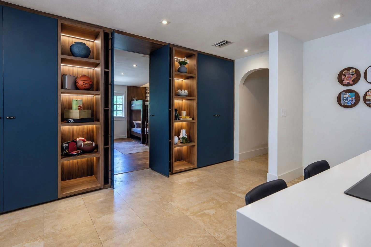 A modern interior with a large open shelving unit containing sports equipment and decorative items, a bunk bed in the background, and a white table with chairs in the foreground.