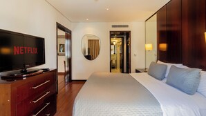Habitación de hotel con una televisión de pantalla plana grande sobre un aparador de madera, una cama con ropa blanca y dos almohadas, un espejo en la pared y una puerta que da a un baño.