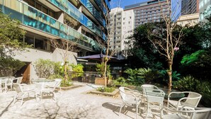 Un espacio al aire libre con sillas y mesas de mimbre, rodeado de follaje y edificios modernos.