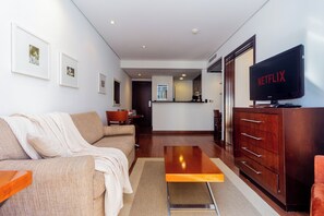 Ein modernes Wohnzimmer mit einer Couch, einem Couchtisch, einem Fernseher und einer kleinen Küche im Hintergrund.