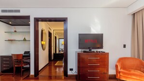 Una habitación de hotel moderna con un aparador de madera, un televisor, un sillón naranja cómodo y un área de comedor con una computadora portátil y libros.
