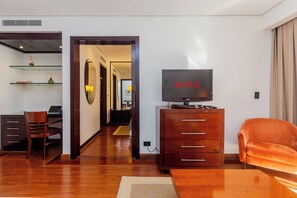 Ein modernes Hotelzimmer mit einem Holzkonsolenmöbel, einem Fernseher, einem bequemen orangefarbenen Sessel und einer Essecke mit einem Laptop und Büchern.