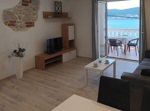 Ein Wohnzimmer mit Fernseher, Couchtisch und Sofa. Es gibt einen Balkon mit Meerblick.