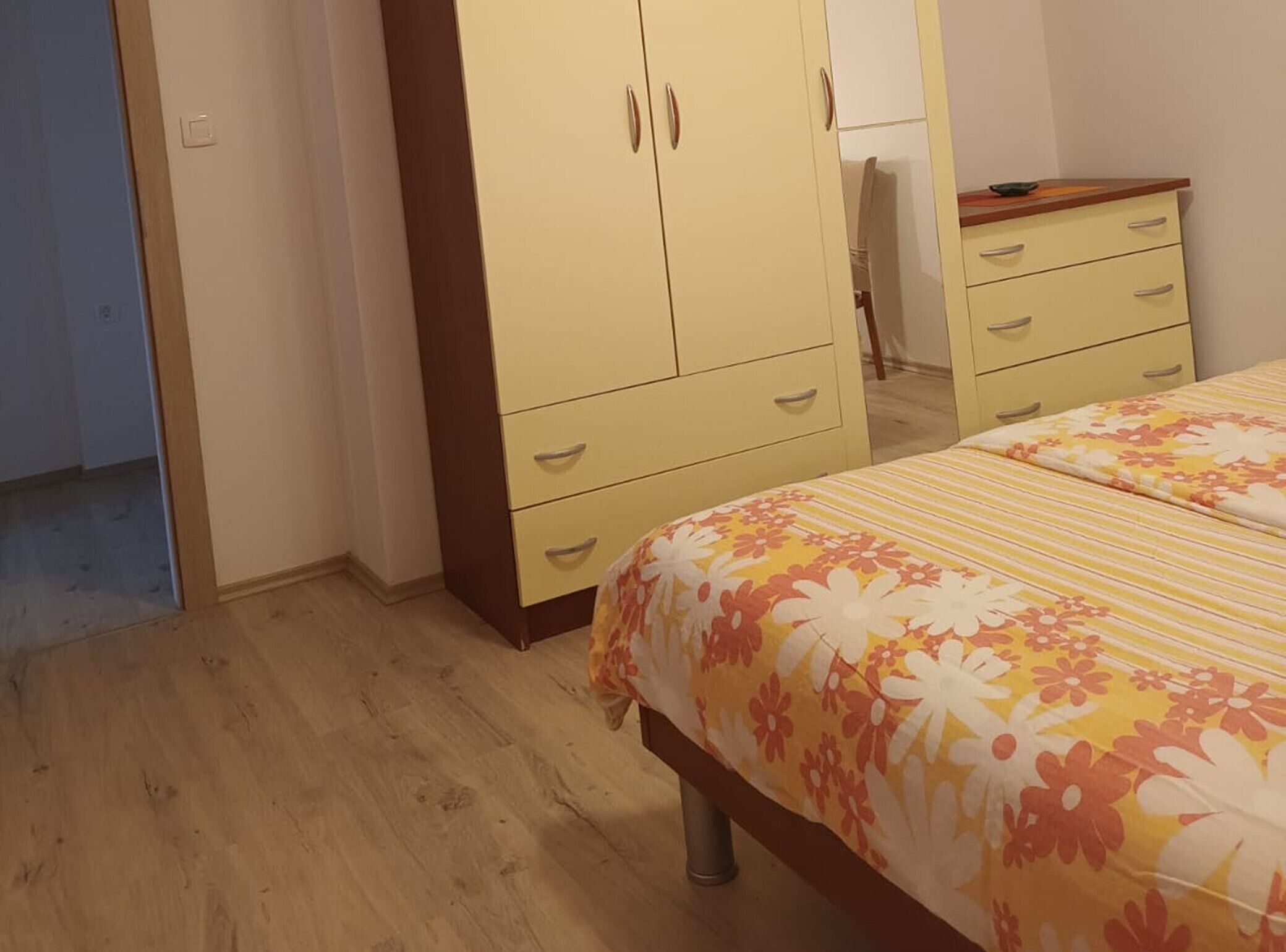 A bedroom with a wooden floor, a bed with floral bedding, a wardrobe with two doors, and a small table with a chair.