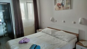 Hotelkamer met een bed, nachtkastje, schilderij, raam met gordijnen en uitzicht op een gang.