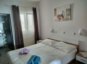 Ein Hotelzimmer mit Bett, Nachttisch, einem Gemälde, einem Fenster mit Vorhängen und Blick auf einen Flur.