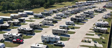 Un grand terrain de camping pour véhicules récréatifs, avec de nombreuses rangées de véhicules et de remorques, des aires de stationnement bien entretenues et un paysage environnant composé d’arbres et de champs ouverts.