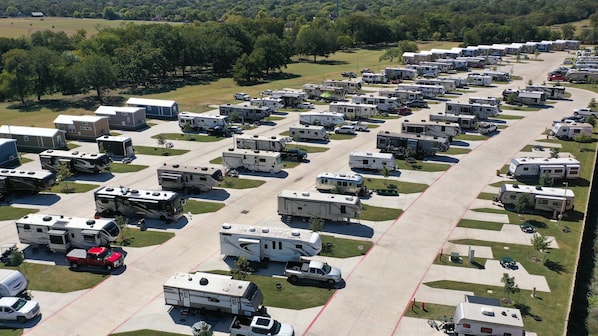 A large RV park with multiple rows of RVs and trailers, well-maintained parking areas, and a surrounding landscape with trees and open fields.