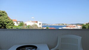 A balcony with a table and a chair, overlooking a coastal town with buildings and a clear sky. - Classic One Bedroom Apartment with Sea View and Balcony (Rogoznica)
