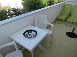 Ein Balkon mit einem kleinen weißen Tisch und zwei weißen Stühlen, einem runden Tisch mit einer schwarzen Ablage sowie zwei grünen Stühlen.
