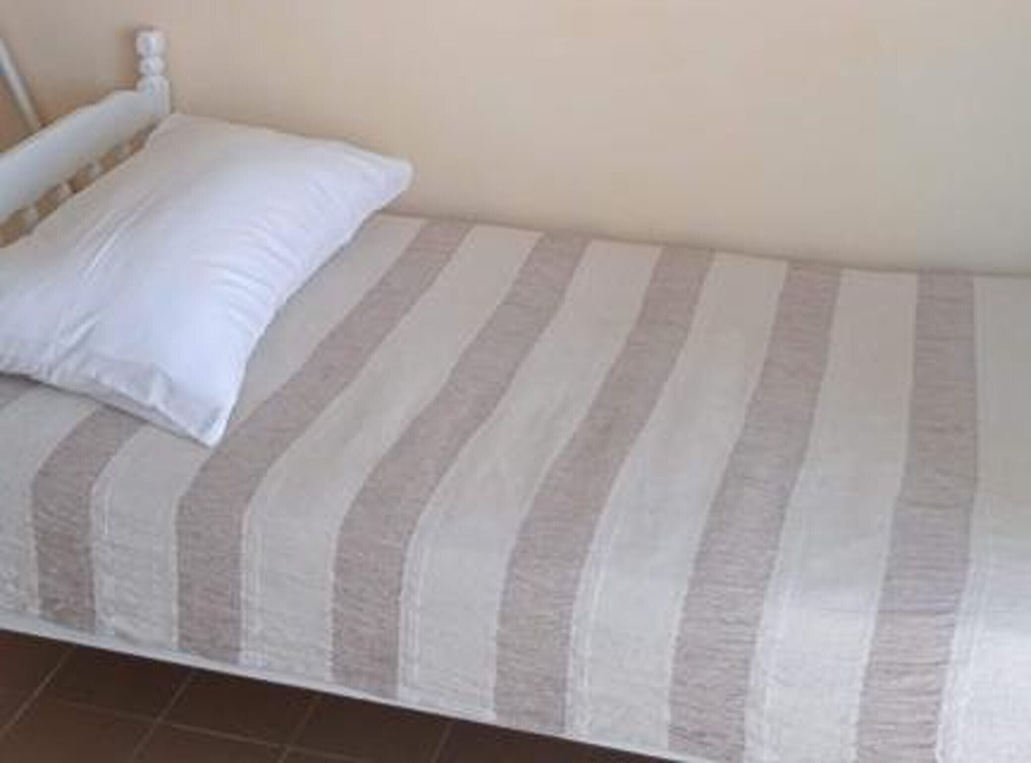 A single bed with a striped bedspread and a white pillow in a room with plain walls and a tiled floor.