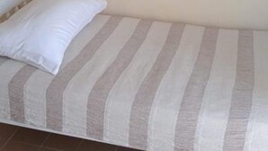 Una cama individual con una colcha a rayas y una almohada blanca en una habitación con paredes lisas y piso de baldosas.