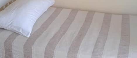 A single bed with a striped bedspread and a white pillow in a room with plain walls and a tiled floor.