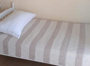 Ein Einzelbett mit gestreifter Bettdecke und einem weißen Kopfkissen in einem Zimmer mit schlichten Wänden und einem gefliesten Boden.