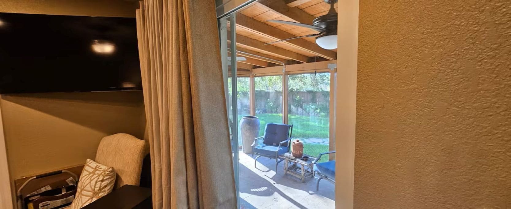 A room with a television mounted on the wall, a glass door leading to an outdoor seating area with a table and chairs, and a ceiling fan.