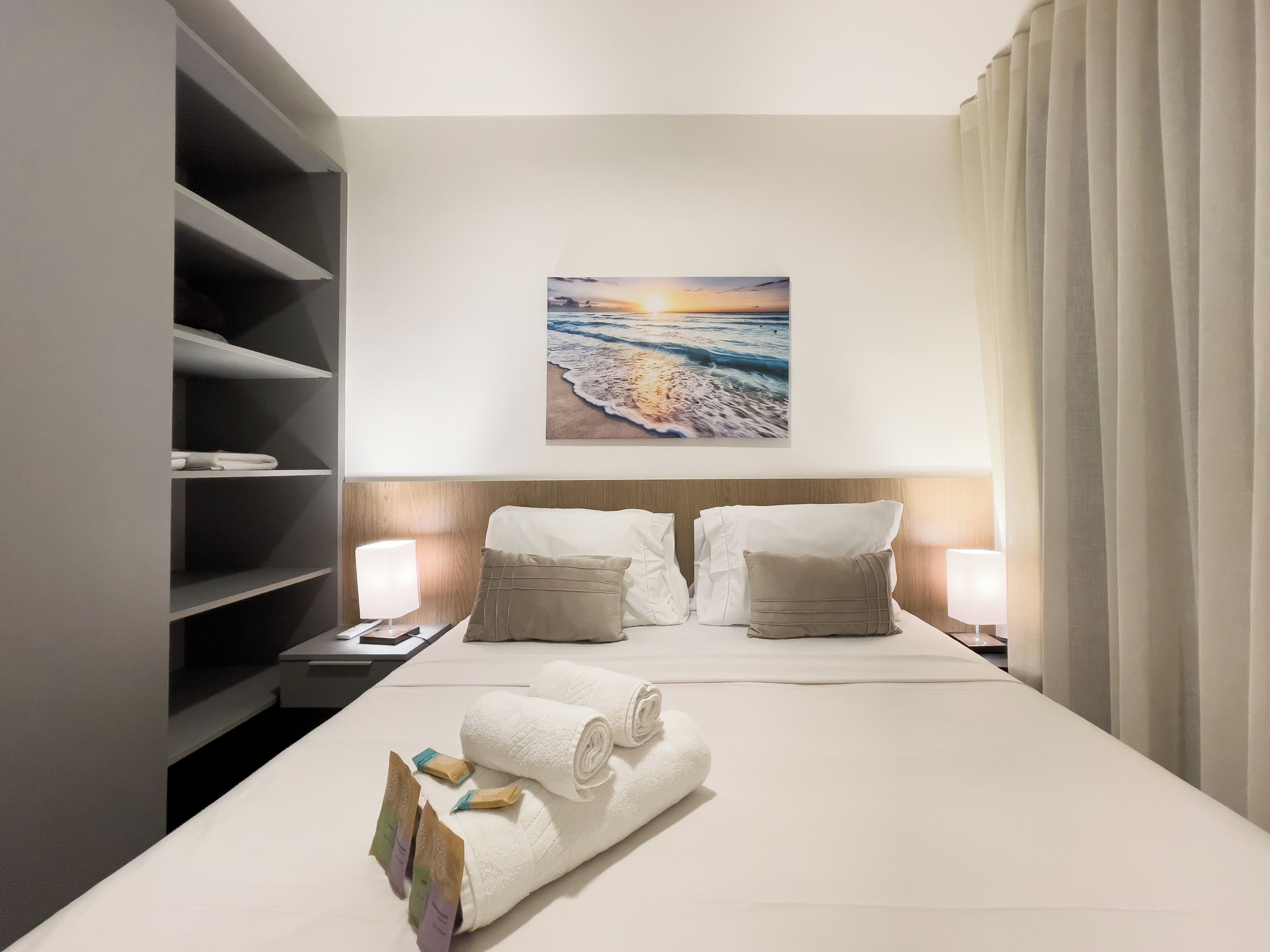 A hotel room with a bed, a nightstand, a wall-mounted painting, and a large window with curtains.