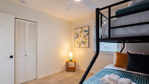 A bunk bed room with a ceiling fan, a door with louvered panels, a small table with a lamp, and a painting on the wall.