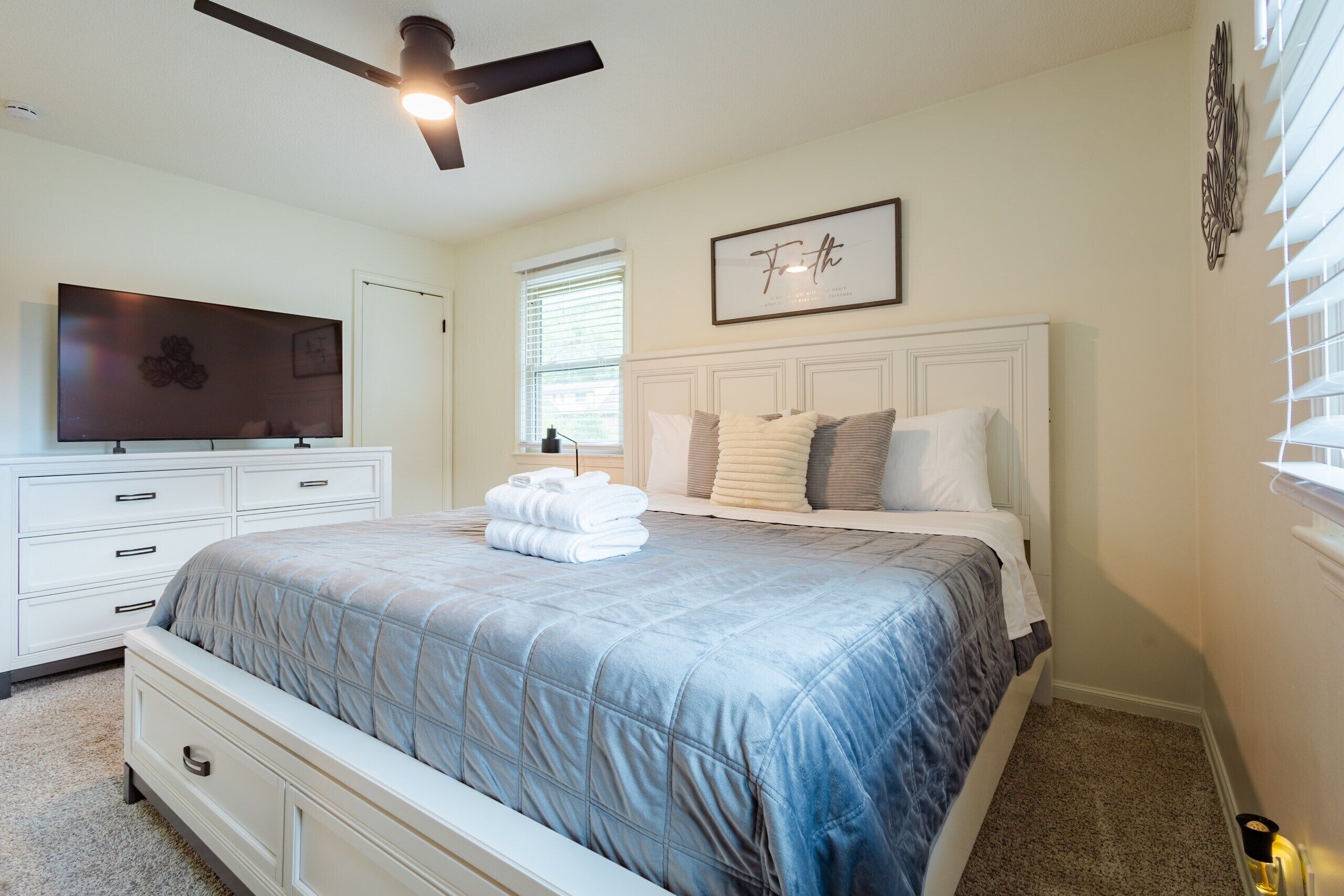 A bedroom with a bed, a television, a ceiling fan, and a framed quote on the wall.