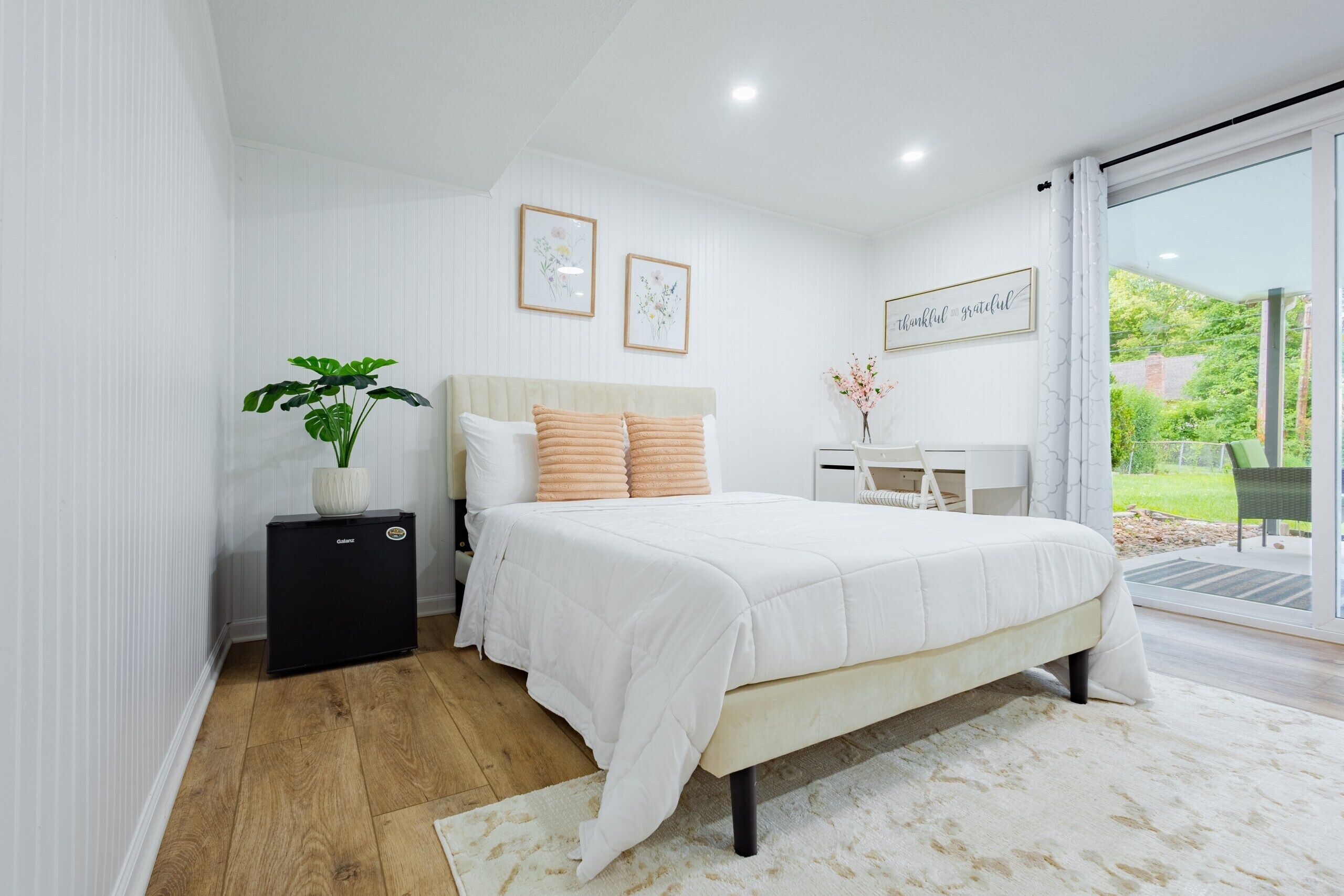 A bedroom with a bed, a bedside table, a potted plant, and a view of the outdoors.