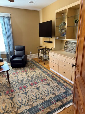 A living room with a black leather chair, a wooden coffee table, a patterned rug, a wooden dresser, a television mounted on the wall, and a window with curtains.