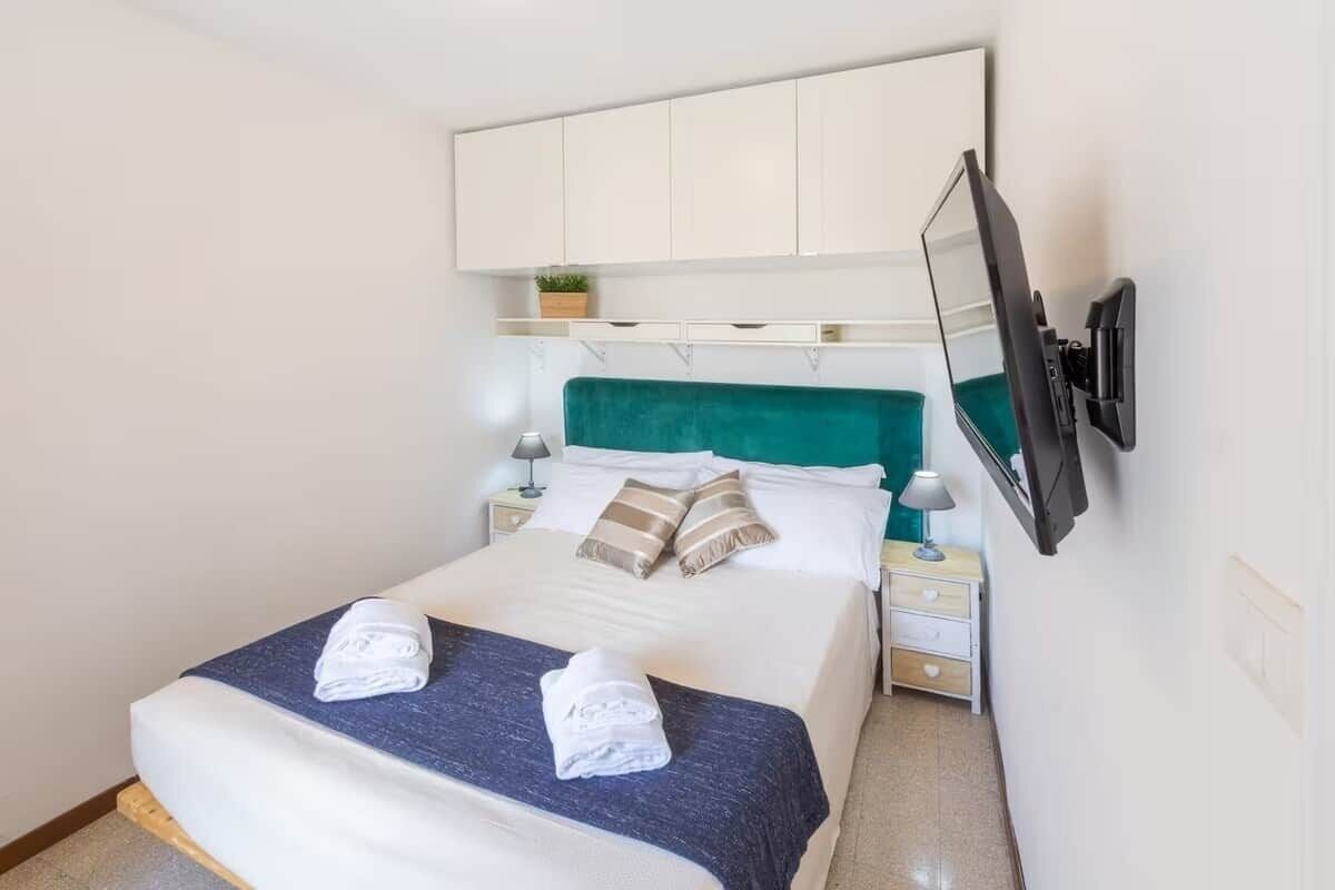 A hotel room with a bed, a television mounted on the wall, and a small bedside table with a lamp.